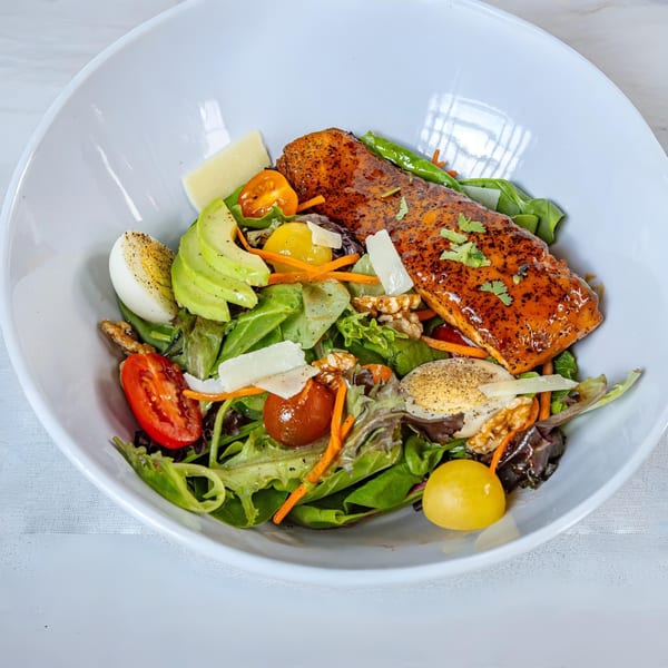 A MiMi's Craft Kitchen salmon salad with avocado, eggs, tomatoes, and greens in a white bowl.